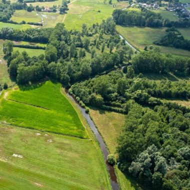 Luchtfoto van akkers en bosgebied in de vallei van de Aa. De Aa zelf slingert zich door het landschap. Op de akkers zijn enkele grote waterplassen zichtbaar.
