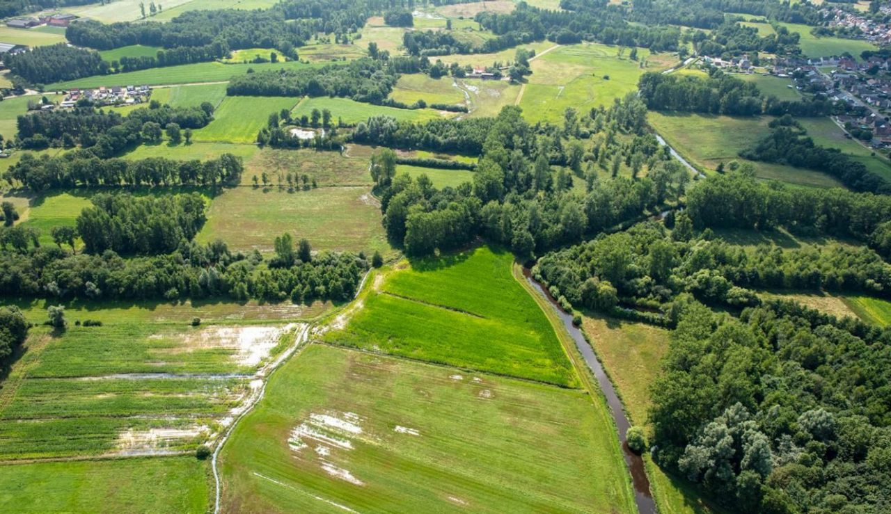 Luchtfoto van akkers en bosgebied in de vallei van de Aa. De Aa zelf slingert zich door het landschap. Op de akkers zijn enkele grote waterplassen zichtbaar.
