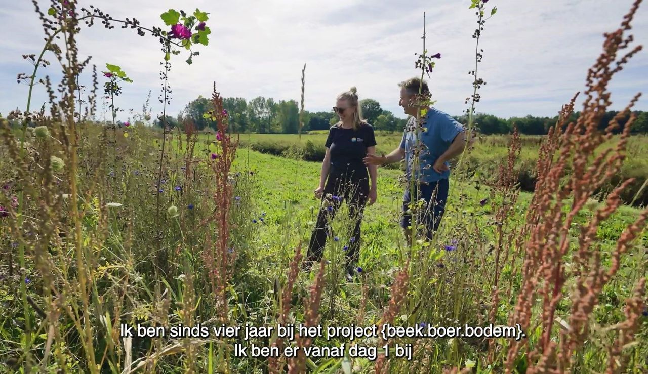 Een vrouw en man staan samen in een groen veld en kijken lachend naar een hoge, bloeiende bloemenrand. Op de achtergrond loopt een gracht tussen de percelen.