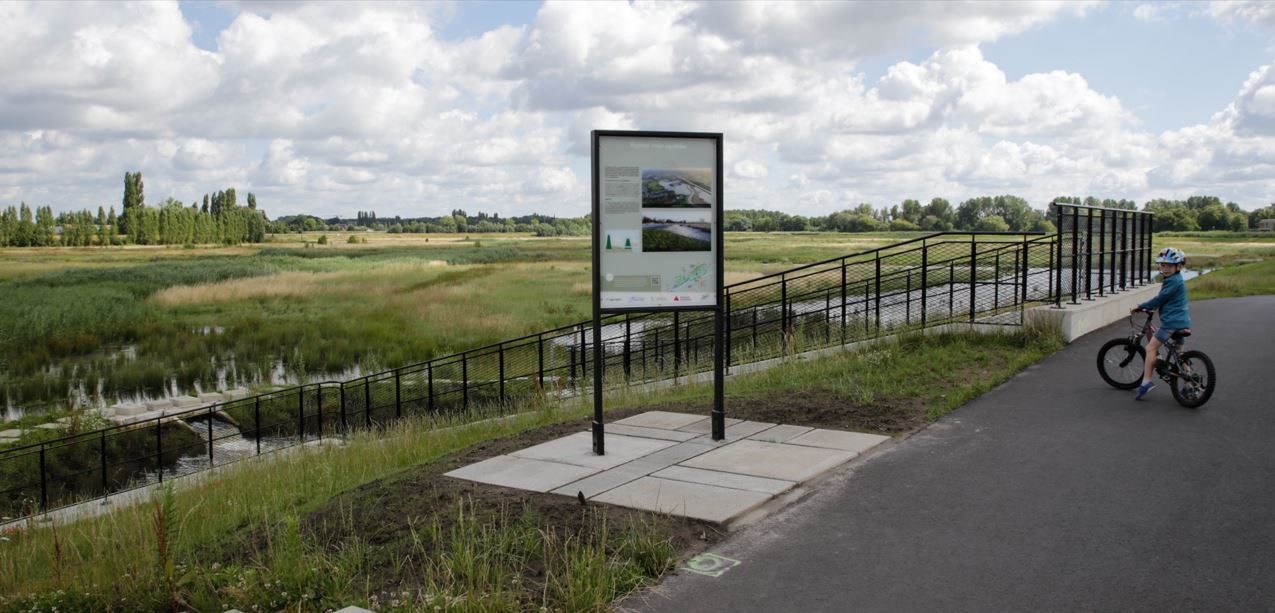 Een jong kind staat met de fiets bij een informatiebord, geplaatst naast de ontwateringssluis van het overstromingsgebied Polder van Lier.