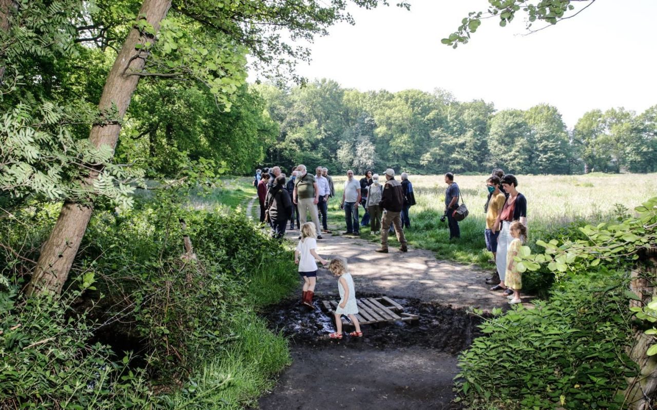 Een groep mensen krijgt uitleg van de boswachter van domein Ertbrugge. Ze staan op een pad, tussen bomen en grasveld. Enkele kinderen spelen in de modder.