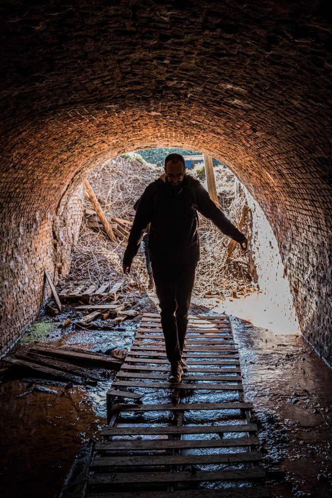 Een persoon loopt over paletten in een gemetselde tunnel. Rondom rond liggen waterplassen, hout- en takkenafval. 