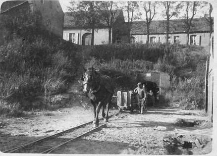 Een oud zwart-wit beeld van een paard dat een rij kleiwagentjes doorheen een tunnel trekt. De karretjes lopen over een smalspoor. Enkele arbeiders begeleiden het transport. 