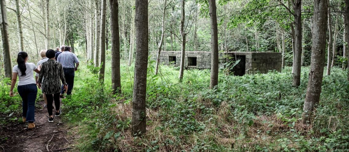 Enkele mensen lopen tussen bomenrijen. Een kleine bunker van betonblokken staat tussen de bomen.