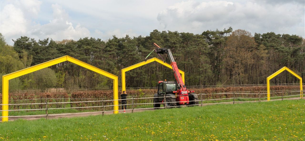 Een hijskraan tilt een grote, stalvormige stalen contour op haar plaats tussen de velden. Links en rechts staan gelijkaardige structuren al op hun plek. 