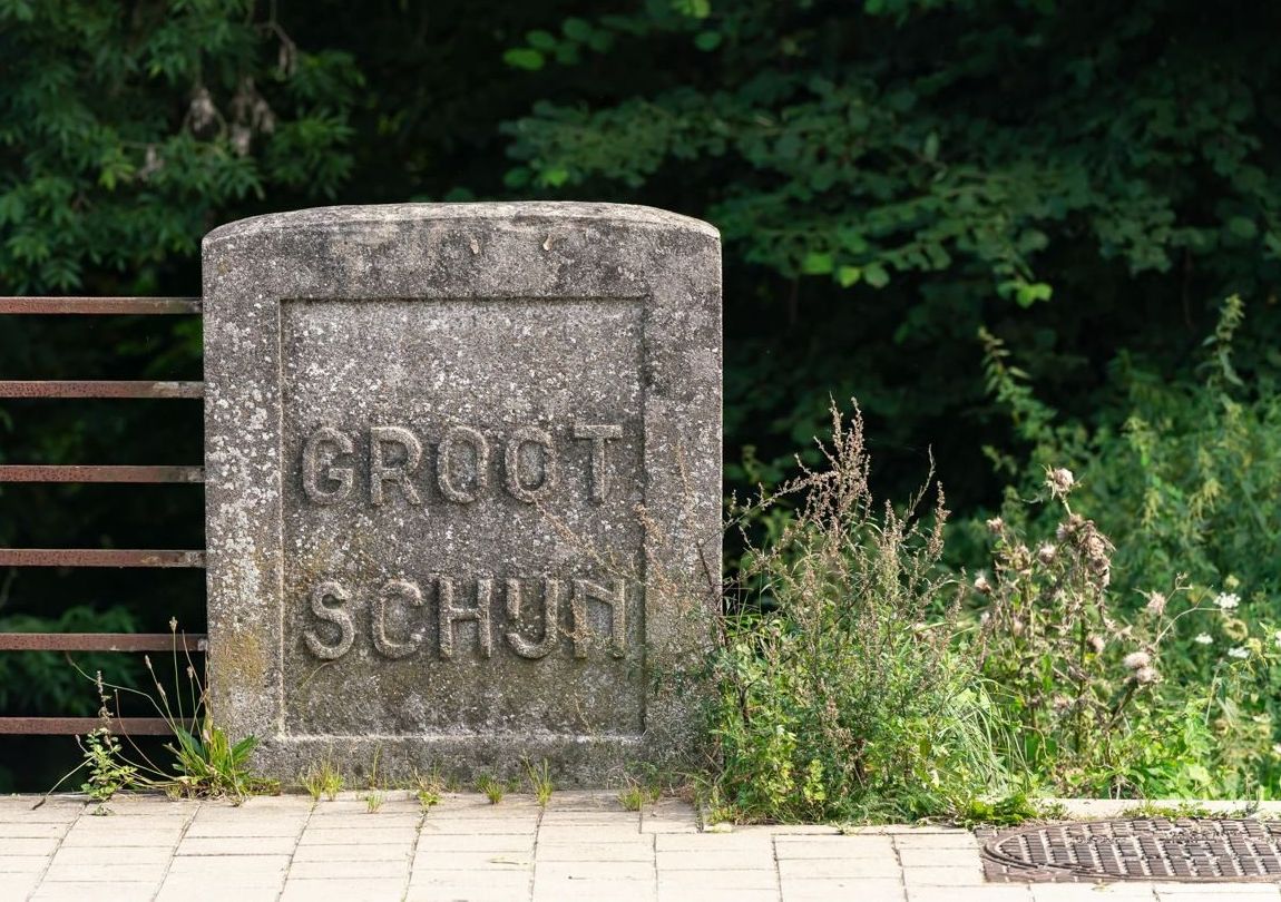 Een oude naamsteen draagt de naam 'Groot Schijn'. De staan staat aan de rand van de stoep, met aan de linkerzijde een roestige leuning. Achter steen en leuning ligt een diepte waar bomen staan.
