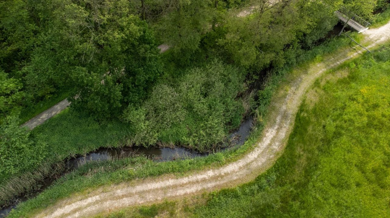 Een smal waterlinkt kronkelt door grasveld, met op de ene oever bomen en struiken. Parallel naast de rivier loopt een zandpad. Een klein brugje ligt over de rivier.