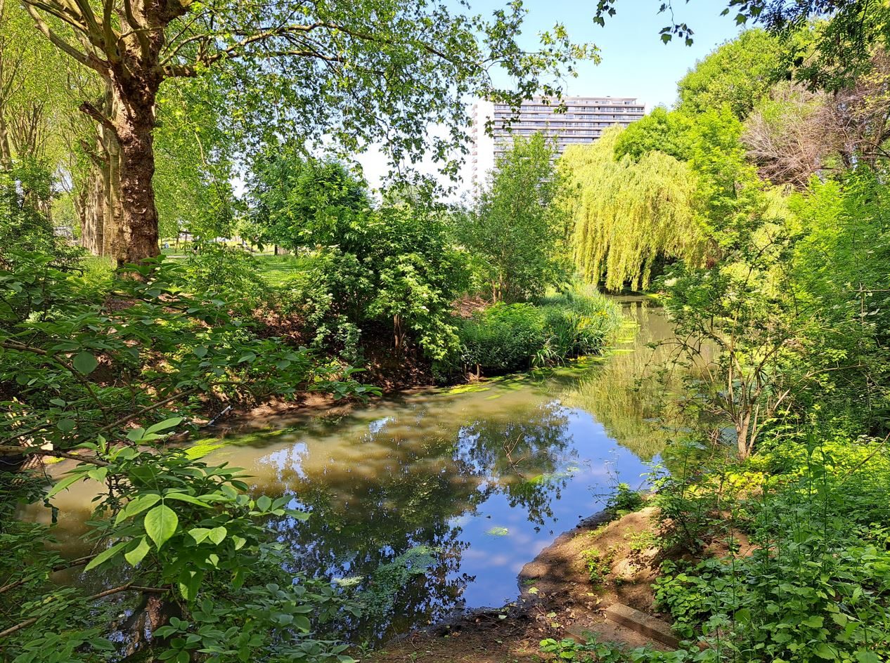 Een rivier stroomt in een parklandschap met bomen en struiken. Op de achtergrond verrijst een hoog flatgebouw.