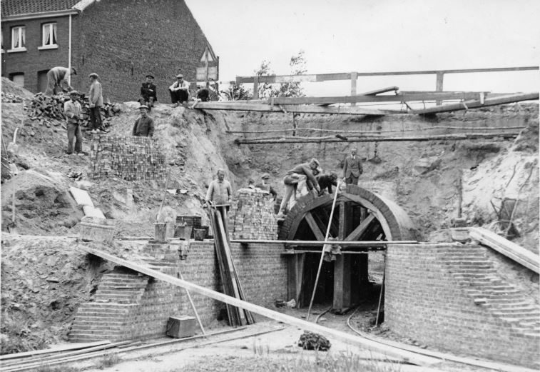 Zwart-wit foto van een tunnel in opbouw. Enkele arbeiders staan bovenop de tunnelboog en leggen bakstenen. Op de helling naast de tunnel staan nog enkele arbeiders bij stapels bakstenen. Vanop de hogergelegen straat kijken enkele mensen gehurkt toe. De straat is bovenaan afgezet met een houten hekwerk. Op de voorgrond liggen planken en ander bouwmateriaal.