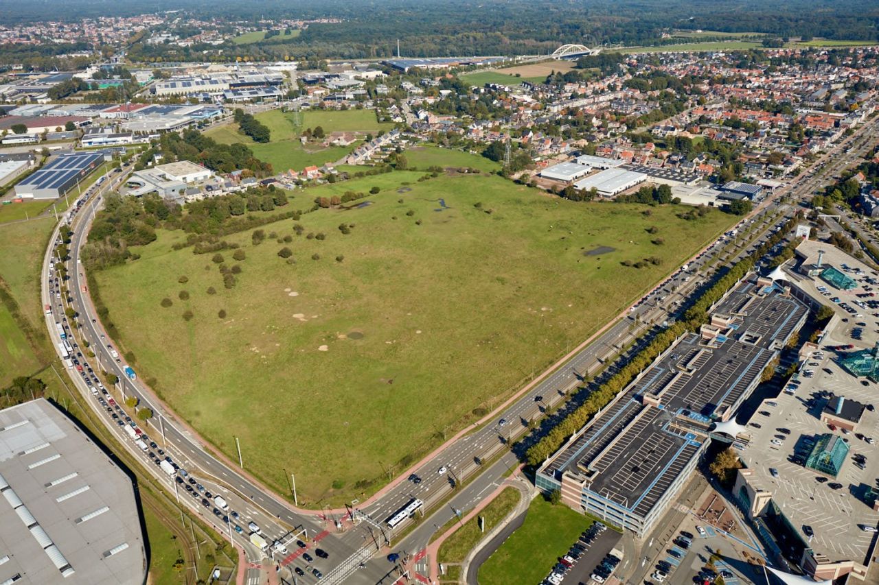 Een groene grasvlakte is aan twee zijden omzoomd met drukke verkeerswegen. Rechts vooraan liggen de gebouwen van het Wijnegem Shopping Center. 