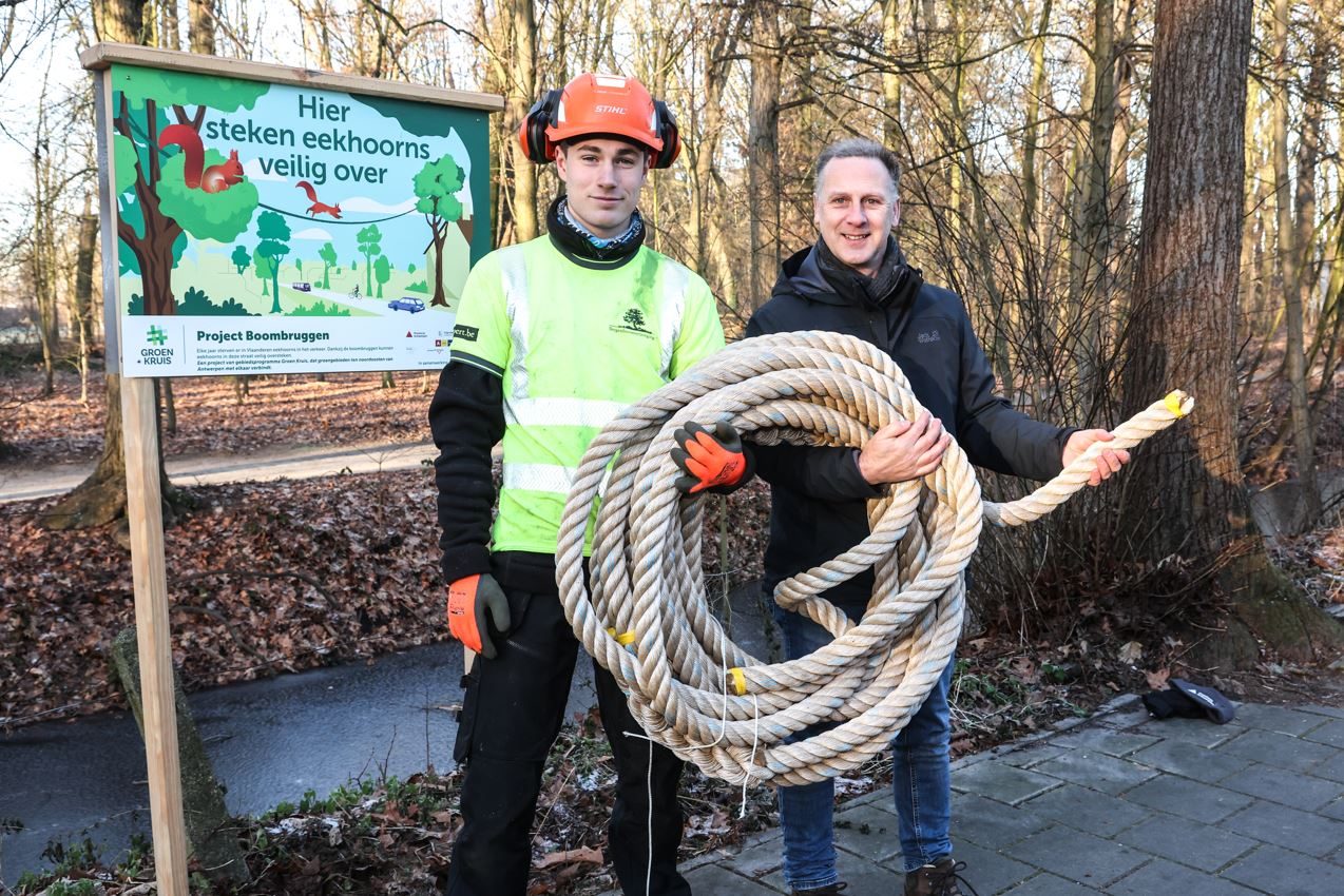 Peter Verdyck, directeur van het provinciaal domein Rivierenhof, en een medewerker poseren bij een infobord over de boombruggen. Samen houden ze een rol  stevig 'boombruggentouw' vast. 