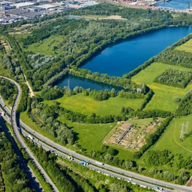 Het natuurgebied Bospolder in Ekeren vanuit de lucht gezien. Een grote en een kleine waterplas liggen omzoomd door groen. Op de achtergrond zi je de Antwerpse haven, vooraan loopt de A12 en aan de rechterzijde zie je het rangeerstation Antwerpen Noord.