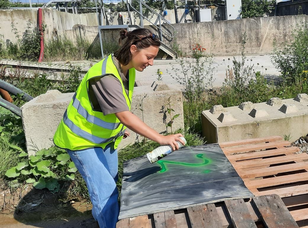 Een persoon staat op een industrieel terrein. Ze buigt zich over een rubberplaat en spuit er met een verfspuit een getal op.
