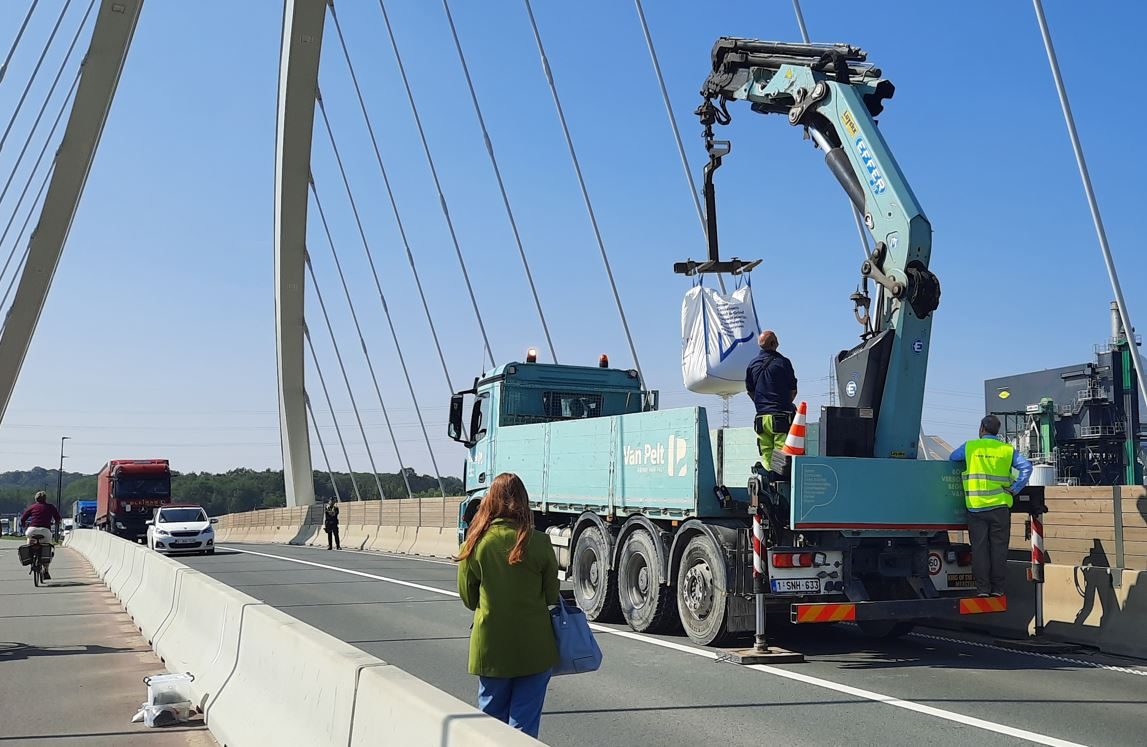 Een vrachtwagen met hefkraan staat op een brugdek. De kraan tilt een grote, zware zak over de omheining naast de rijstroken. Enkele personen staan rondom.