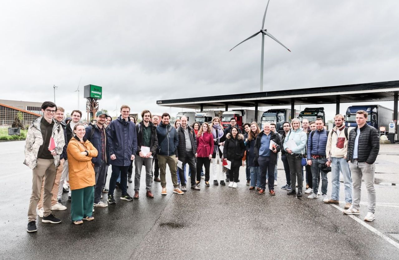 Een groep van ongeveer dertig mensen poseert lachend voor de camera. Ze staan op een natgeregend astfalt ondergrond. Achter hen staat een rij vrachtwagens die brandstof tanken bij een benzinestation. In de achtergrond is een moderne windmolen met drie wieken zichtbaar. 