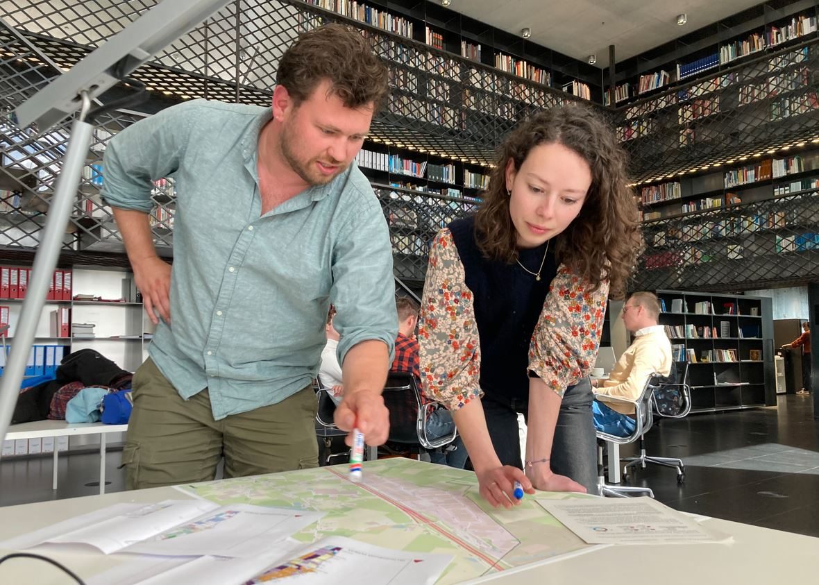 Twee personen staan gebogen over een tafel, waarover kaartmateriaal en andere documenten zijn uitgespreid. Achter hen zijn nog andere mensen zichtbaar die rond een tafel zitten. Op de achtergrond zie je drie verdiepingen stalen boekenrekken, gevuld met boeken, en met een gangpad eromheen.