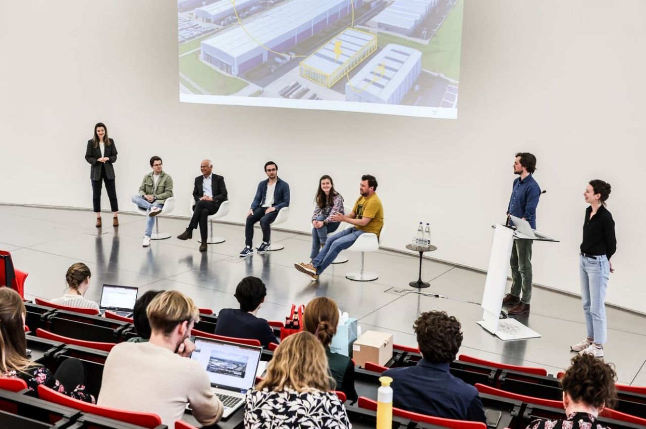 Op een metalen podium zitten en staan in totaal 8 mensen. Op het scherm achter hen is een deel van een fotopresentatie zichtbaar. Deze toont enkele gebouwen waarover met een kleurstift andere posities/gebouwvormen zijn ingetekend. Op de voorgrond van de foto zie je mensen vanop de rug; zij zitten op rijen stoelen en kijken naar de presentatie op het podium. 
