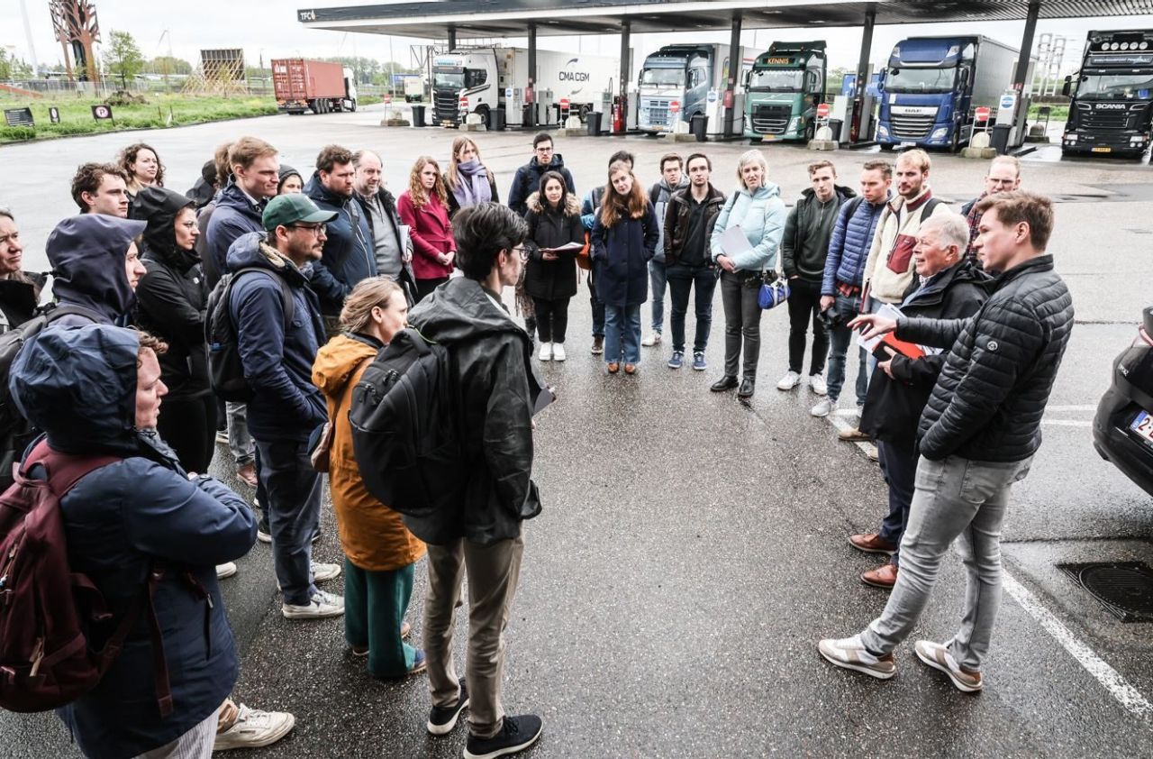 Een groep van een dertigtal mensen staat in een kring buiten op het asfalt. Achter hen is een rij vrachtwagens zichtbaar die brandstof tanken bij een benzinestation.