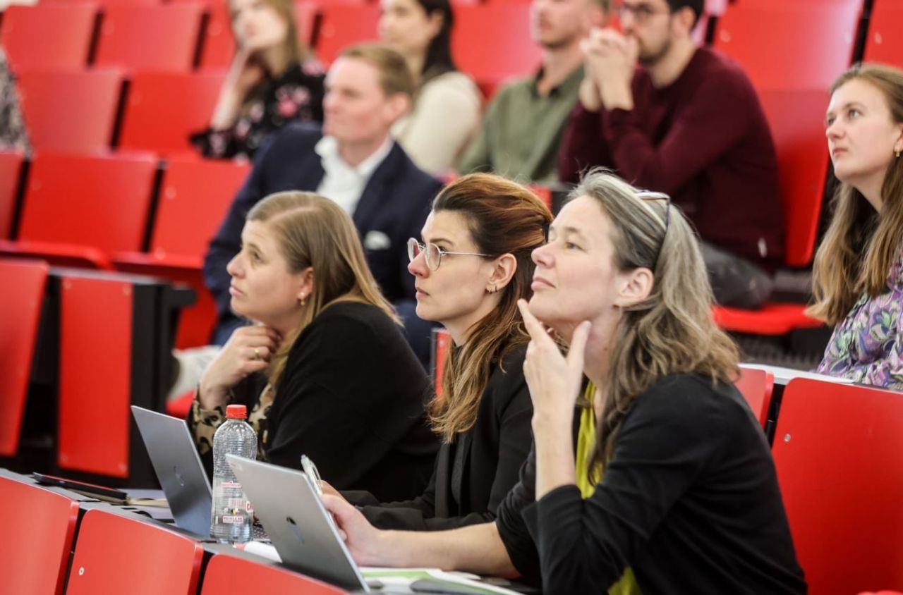 Meerdere mensen zitten op rijen rode stoelen. Ze hebben een laptop voor zich en kijken aandachtig naar iets wat buiten beeld is (een presentatie).