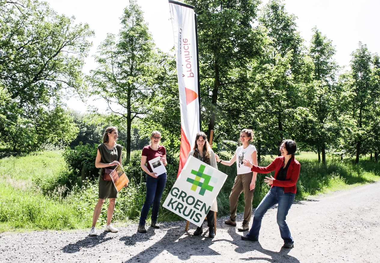Vijf personen staan op een landweg voor een groepje bomen. Achter hen staat een strandvlag met het logo van de provincie Antwerpen. De middelste persoon draagt een groot bord met het logo van Groen Kruis.