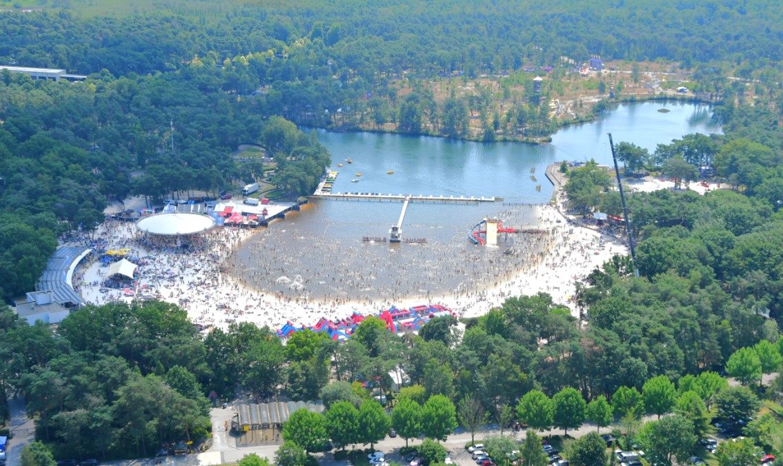 Luchtfoto: tussen de bomen ligt een meer met een strand. Tal van strand- en zwemvoorzieningen zijn zichtbaar (redderstoren, glijbanen, aanlegsteiger,...) Heel veel bezoekers zijn als kleine stipjes zichtbaar op het strand en in het water.