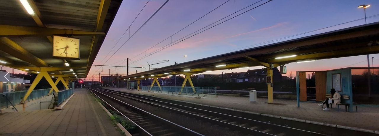 Sfeerbeeld van het perrons en de sporen van het station van Boom bij valavond. De hemel kleurt blauw-roze. Er zijn geen treinen, slechts een persoon zit te wachten op een bank.
