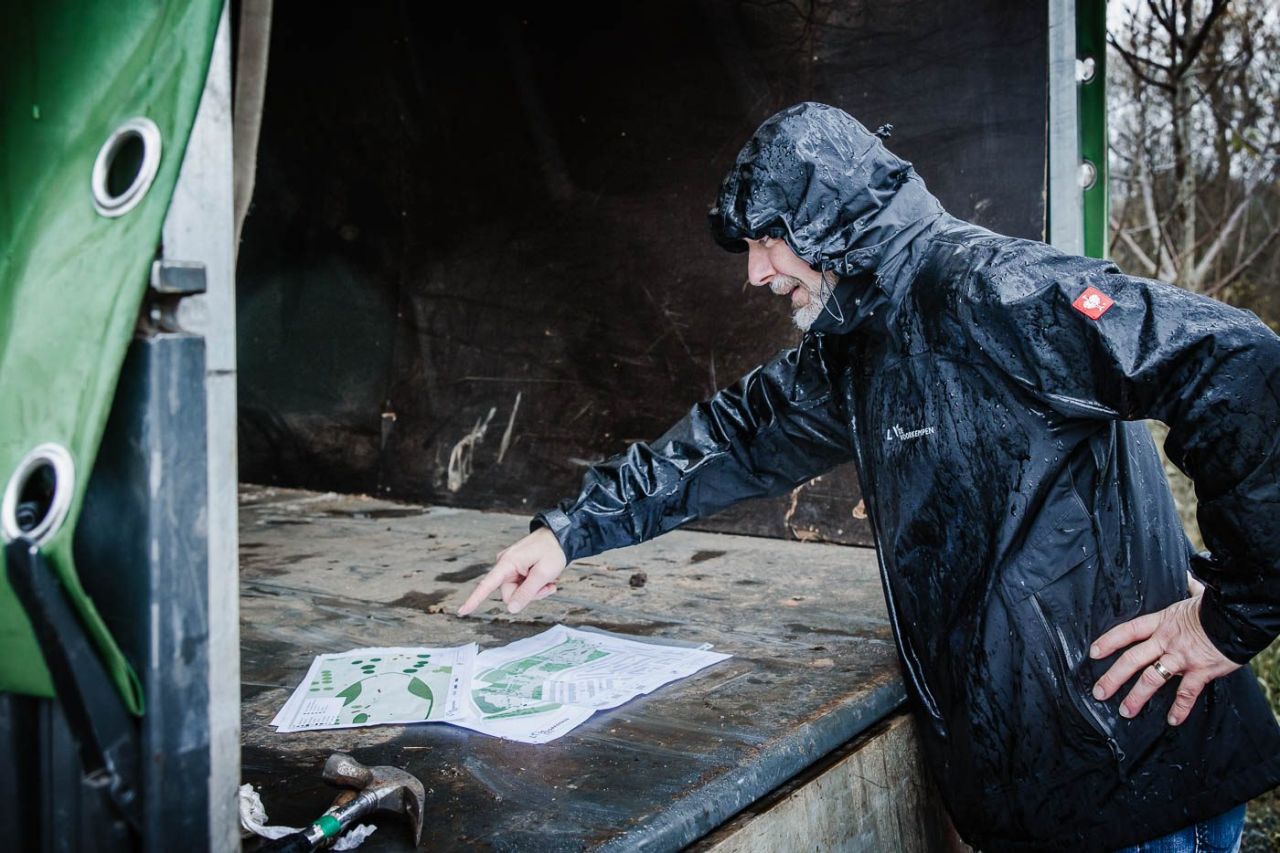 Een man in doorweekte regenjas staat bij de open laadruimte van een vrachtwagen. Hij kijkt en wijst naar enkele ontwerpplannen die in de laadruimte liggen.