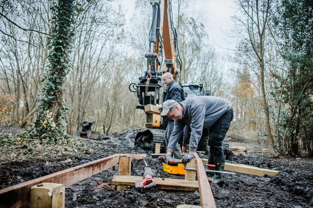Een arbeider staat diep in de modder en buigt zich over de houten, halfafgewerkte constructie van een vlonderpad. Achter hem doemt een grote graafkraan op.