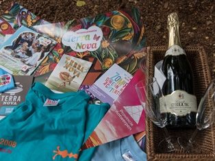 Close-up van een verzameling drukwerk en t-shirts uit het archief van Terra Nova, ernaast een mandje met een fles schuimwijn en twee glazen, op de achtergrond een droog bladerdek in de tuin van Arboretum Kalmthout.