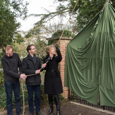 Plechtige inhuldiging van een toegangshek in Arboretum Kalmthout. Twee mannen en een vrouw trekken aan een touw het groene zeil omhoog, dat over het hek hangt.