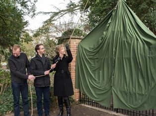Plechtige inhuldiging van een toegangshek in Arboretum Kalmthout. Twee mannen en een vrouw trekken aan een touw het groene zeil omhoog, dat over het hek hangt.