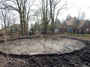 Een tuinier plant jonge haagstruiken aan in een grote cirkel op vers omgespitte aarde. Op de achergrond de cottage in Arboretum Kalmthout.
