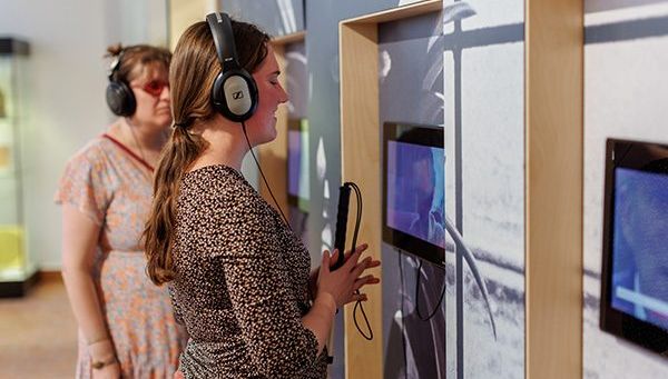 In het Gasthuismuseum Geel hangen drie schermen aan de muur, waaraan koptelefoons verbonden zijn via kabels. Een blinde en een slechtziende vrouw hebben allebei zo’n koptelefoon op en beluisteren het filmpje dat afspeelt op de schermen.