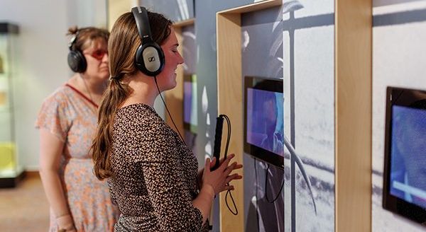 In het Gasthuismuseum Geel hangen drie schermen aan de muur, waaraan koptelefoons verbonden zijn via kabels. Een blinde en een slechtziende vrouw hebben allebei zo’n koptelefoon op en beluisteren het filmpje dat afspeelt op de schermen.