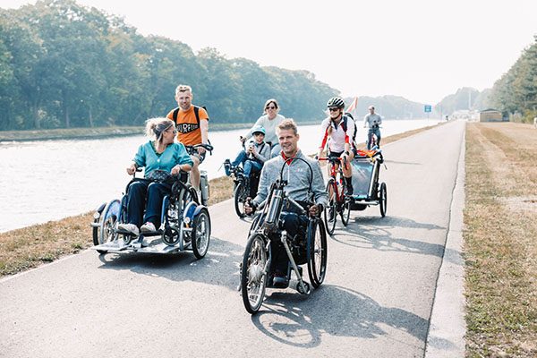 Een groepje van zeven personen fietst op het jaagpad langs het kanaal Dessel Schoten in Mol. Ze zitten elk op een toegankelijke fiets: de eerste man beweegt zich voort met een handbike, een vrouw zit op een rolstoelfiets die wordt bestuurd door haar echtgenoot. Daarachter fietst een vrouw met haar zoon in een semi-ligfiets tandem. Een andere vrouw die achterop fietst, draagt een helm en heeft een fietskar achter aan haar fiets bevestigd. De zon schijnt.