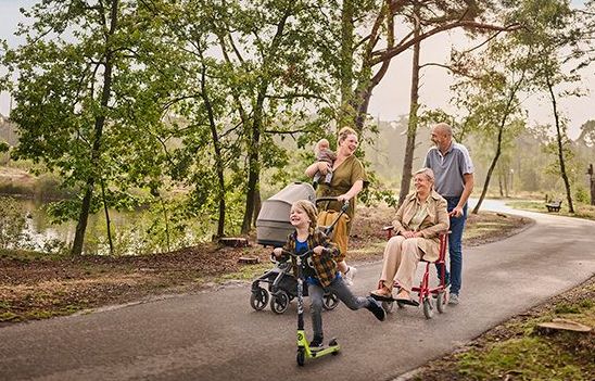 Een moeder wandelt met haar twee kinderen op een verharde ondergrond tussen de bomen aan het water. Eén van haar kinderen is nog een baby en houdt ze in haar rechterarm, terwijl ze met haar linkerarm een buggy duwt. Het oudere zoontje gaat snel vooruit op zijn step. Naast dit drietal wandelt een ouder koppel: de vrouw zit in een rolstoel, die wordt voortgeduwd door haar echtgenoot.
