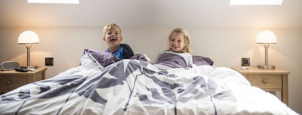 In een hotelkamer liggen twee kinderen – steunend op hun ellebogen – te lachen in het grote tweepersoonsbed van de ouders. Aan weerszijden van het bed staan twee nachtkastjes met daarop een brandend nachtlampje, ook al schijnt er zonlicht door de twee dakramen.