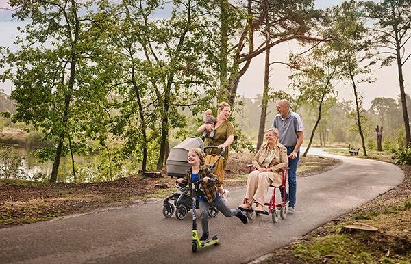 Een moeder wandelt met haar twee kinderen op een verharde ondergrond tussen de bomen aan het water. Eén van haar kinderen is nog een baby en houdt ze in haar rechterarm, terwijl ze met haar linkerarm een buggy duwt. Het oudere zoontje gaat snel vooruit op zijn step. Naast dit drietal wandelt een ouder koppel: de vrouw zit in een rolstoel, die wordt voortgeduwd door haar echtgenoot.