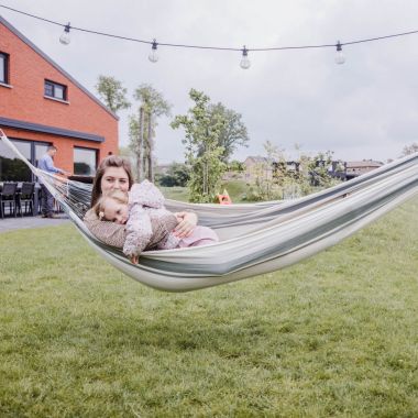 Bij vakantiewoning Den Akker in Mol ligt een moeder samen met haar jonge dochter in een stoffen hangmat. Ze genieten van de rust op het grasveldje in de landelijke omgeving. Op de achtergrond barbecuet de vader op het betegelde terras. Naast hem staat een lange tuintafel met lege stoelen.