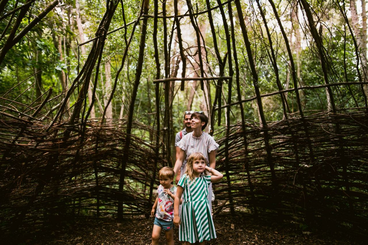 Een gezin met twee jonge kinderen gaat een wilgenhut in het Trollenbos van De Schorre in Boom binnen. Het wilgenriet is niet aansluitend, waardoor er licht binnenkomt, voornamelijk langs de bovenkant. De vrouw kijkt naar de dakconstructie boven haar dat licht doorlaat. Ze moedigt de kinderen met een klein duwtje in de rug aan om verder te wandelen, terwijl haar man achter haar volgt. De kinderen kijken vol bewondering rondom zich.
