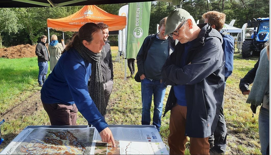 Een sfeerfoto van de demomarkt {beek.boer.bodem} van oktober 2021, buiten in een veld. Een persoon wijst naar een poster met luchtfoto's en grafieken, hij richt zich tot een aandachtige toeschouwer. In de achtergrond staan meerdere kleine groepjes mensen met elkaar te praten in een veld. met tentjes en beachvlaggen.