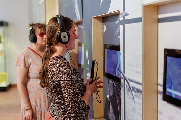 In het Gasthuismuseum Geel hangen drie schermen aan de muur, waaraan koptelefoons verbonden zijn via kabels. Een blinde en een slechtziende vrouw hebben allebei zo’n koptelefoon op en beluisteren het filmpje dat afspeelt op de schermen. 