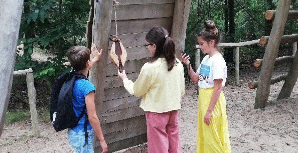 Drie kinderen ontdekken in provinciaal domein Hoge Mouw het bosspel bij een houten speeltuig op een zanderige ondergrond. De jongen leunt tegen het speeltuig en draagt de rugzak met de spelattributen, terwijl het meisje rechts het gps-toestel bedient. Een ander meisje staat in het midden en houdt een aanwijzing van het bosspel vast in haar linkerhand.