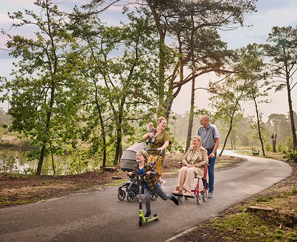 Een moeder wandelt met haar twee kinderen op een verharde ondergrond tussen de bomen aan het water. Eén van haar kinderen is nog een baby en houdt ze in haar rechterarm, terwijl ze met haar linkerarm een buggy duwt. Het oudere zoontje gaat snel vooruit op zijn step. Naast dit drietal wandelt een ouder koppel: de vrouw zit in een rolstoel, die wordt voortgeduwd door haar echtgenoot.