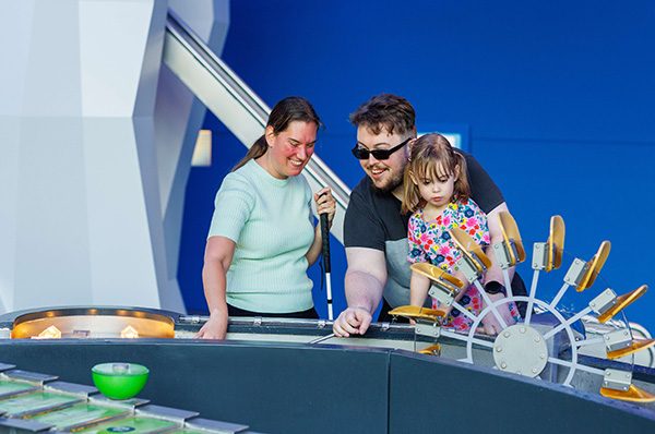 Een mama met taststok, papa met donkere bril en dochter, spelend in Hidrodoe.