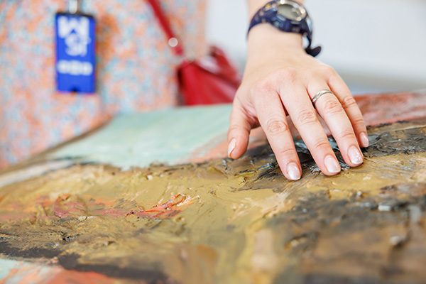 Een close-up van een hand die streelt over een schilderij in het Jakob Smits-museum. Het schilderij ligt horizontaal en bestaat uit dikke lagen verf en bruuske penseelstreken in donkere kleuren. De hand bevoelt het reliëf van de verf. 