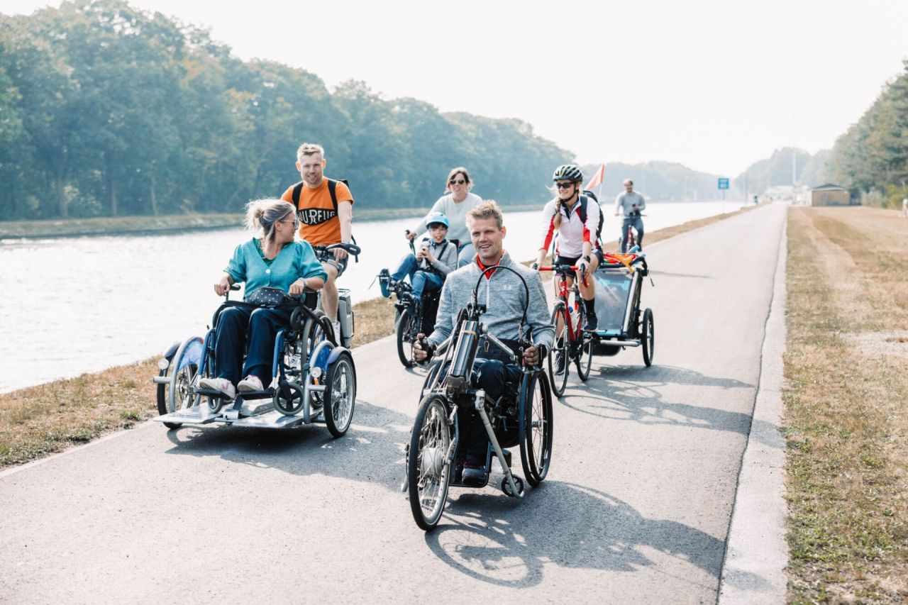 Een groepje van zeven personen fietst op het jaagpad langs het kanaal Dessel Schoten in Mol. Ze zitten elk op een toegankelijke fiets: de eerste man beweegt zich voort met een handbike, een vrouw zit op een rolstoelfiets die wordt bestuurd door haar echtgenoot. Daarachter fietst een vrouw met haar zoon in een semi-ligfiets tandem. Een andere vrouw die achterop fietst, draagt een helm en heeft een fietskar achter aan haar fiets bevestigd. De zon schijnt.