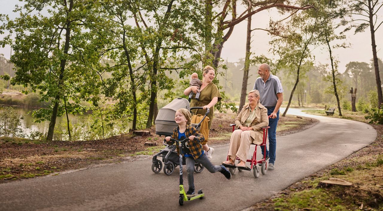 Een moeder wandelt met haar twee kinderen op een verharde ondergrond tussen de bomen aan het water. Eén van haar kinderen is nog een baby en houdt ze in haar rechterarm, terwijl ze met haar linkerarm een buggy duwt. Het oudere zoontje gaat snel vooruit op zijn step. Naast dit drietal wandelt een ouder koppel: de vrouw zit in een rolstoel, die wordt voortgeduwd door haar echtgenoot. 